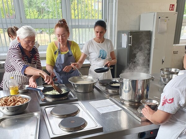 Teilnehmerinnen stehen um den großen Herd und bereiten gemeinsam Speisen zu. Es wird gerührt und es dampft aus den Kochtöpfen. 