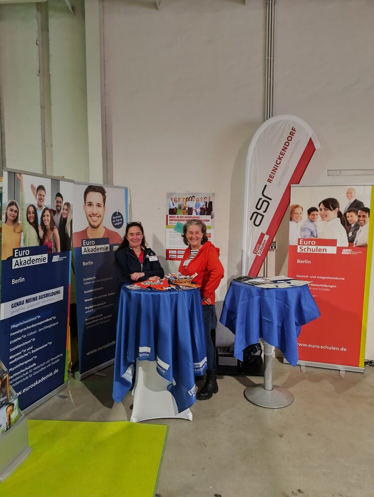 Zwei Frauen, eine in dunkelblau und eine in rot gekleidet, stehen am Messestand der Euro Akademie und Euro-Schulen Berlin und lächeln in die Kamera.