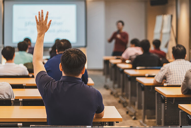 Sommerpause? Nein danke! In einem Unterrichtsraum sitzen verschiedene asiatisch aussehende Personen an Tischen, auf der linken Seite meldet sich eine Person. Ganz vorne ist eine leuchtende digitale Tafel und eine Lehrkraft mit einem Stift in der Hand.