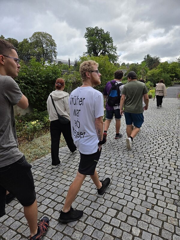 Eine Gruppe von Personen geht auf einem gepflasterten Weg durch einen Park. Im Hintergrund sind Bäume und ein Zaun zu sehen.