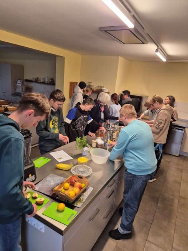 Eine Gruppe junger Menschen arbeitet gemeinsam in einer Küche an verschiedenen Kochaufgaben. Einige schneiden Obst auf Schneidebrettern, während andere Rezepte studieren oder Zutaten vorbereiten. Die Küche ist gut ausgestattet mit modernen Geräten und hell beleuchtet. Die Atmosphäre wirkt konzentriert und gemeinschaftlich.
