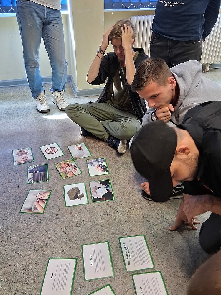 Mehrere junge Erwachsene sitzen und hocken auf dem Boden eines Klassenraums und betrachten grüne Karten mit Texten und Bildern. Einer der Teilnehmer hält sich nachdenklich den Kopf, während andere konzentriert die Karten studieren.