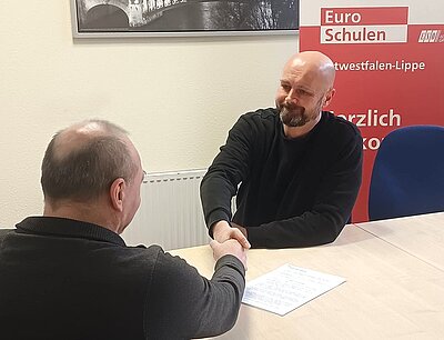 An einem Tisch sitzen ein Mitarbeitender der Euro-Schulen sowie der Teilnehmer Herr B. und geben sich die Hand. Im Hintergrund steht ein großes Roll-up der Euro-Schulen.