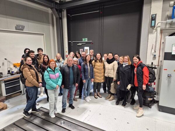 Eine weitere Gruppe von Besuchern posiert für ein Gruppenfoto in einem industriellen oder wissenschaftlichen Labor. Im Hintergrund sind Maschinen und technische Geräte zu sehen.