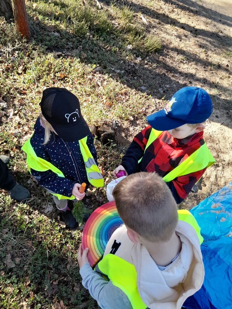 Die Kinder, von denen zwei eine Baseballkappe tragen präsentieren sich gegenseitig offenbar die Ostergeschenke, die sie im Wald gefunden haben. Der Junge ohne Kappe hält einen bunten Regenbogen in der Hand. Die anderen Geschenk kann man schlecht erkennen, da das Bild von oben aufgenommen wurde und die Köpfe sie verdecken.