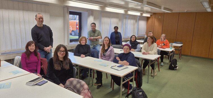 Gruppe von Teilnehmenden in einem hellen Seminarraum: Menschen sitzen und stehen an Tischen mit Unterlagen und Taschen, Blick in die Kamera.