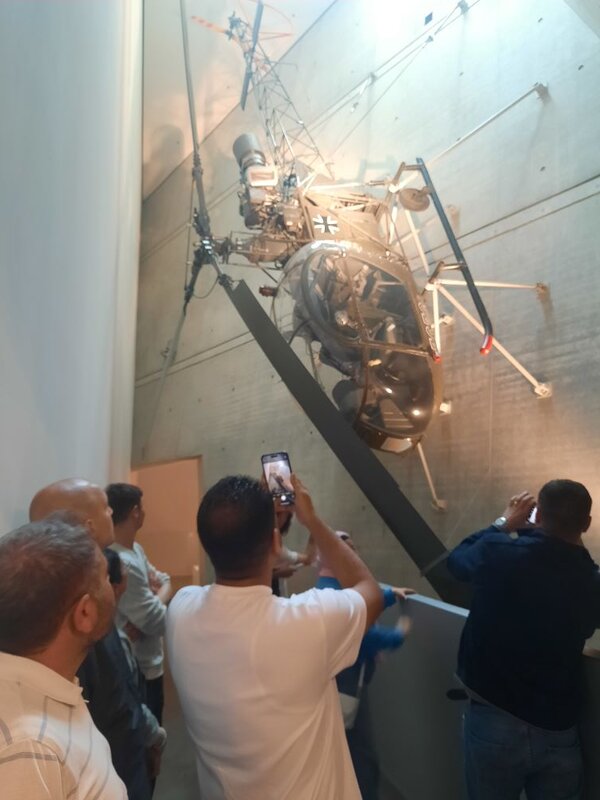 Ein Blick von unten auf eine Gruppe von Menschen, die einen an einer Wand hängenden Militärhubschrauber in einem Museum betrachten. Einige Personen machen Fotos des Exponats.