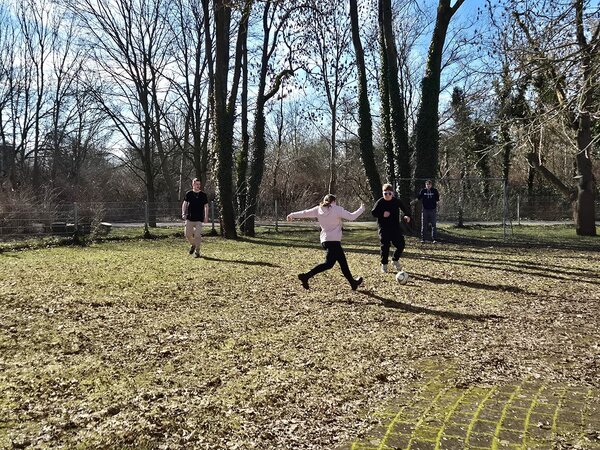 Auf einer Wiese mit Laub tritt eine Teilnehmerin gegen einen Fußball; mehrere Personen stehen in der Nähe, im Hintergrund sind Bäume und ein Torrahmen zu sehen.