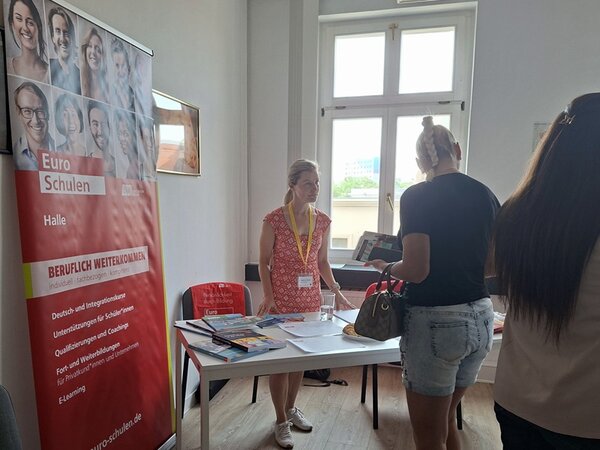 Ein heller Raum mit einem großen Fenster im Hintergrund. Links steht ein rot-weißer Roll-Up mit dem Logo und Namen Euro-Schulen sowie weiteren Informationen. Eine Mitarbeitende der Euro-Schulen mit Namensschild und gelbem Band steht hinter einem weißen Tisch, auf dem verschiedene Broschüren und Papiere liegen. Zwei Besucherinnen stehen vor dem Tisch und informieren sich, eine davon hält eine Handtasche.