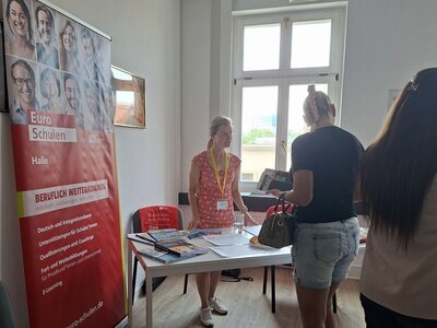 Ein heller Raum mit einem großen Fenster im Hintergrund. Links steht ein rot-weißer Roll-Up mit dem Logo und Namen Euro-Schulen sowie weiteren Informationen. Eine Mitarbeitende der Euro-Schulen mit Namensschild und gelbem Band steht hinter einem weißen Tisch, auf dem verschiedene Broschüren und Papiere liegen. Zwei Besucherinnen stehen vor dem Tisch und informieren sich, eine davon hält eine Handtasche.