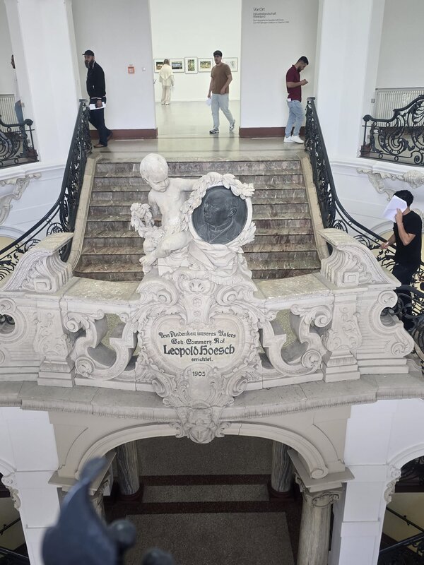 Blick ins helle Treppenhaus des Leopold-Hoesch-Museums: Vor einer breiten Marmortreppe steht eine reich verzierte Steinbalustrade mit Putto und Medaillon, im Hintergrund gehen Menschen durch den Ausstellungsbereich.