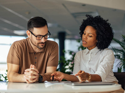 Zwei Personen sitzen an einem Tisch in einem modernen Büro und sprechen miteinander. Rechts sitzt eine Frau mit lockigem Haar und weißer, gestreifter Bluse. Sie zeigt mit einem Stift auf ein Dokument, während der Mann links von ihr mit Brille und braunem Poloshirt aufmerksam zuhört.