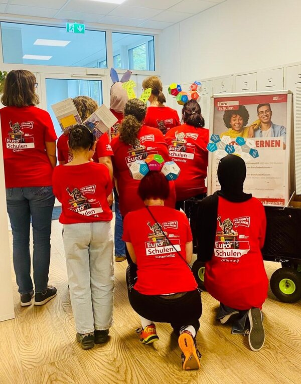In einem Schulflur ist eine Gruppe Menschen von hinten zu sehen. Sie tragen rote Shirts mit dem Logo der Euro-Schulen und zum Teil selbst gebastelten Kopfschmuck. Neben der Gruppe steht ein kleiner Bollerwagen mit einem Poster zum Thema "Deutsch lernen".