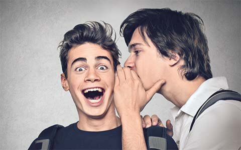 Ein junger Mann flüstert einem anderen jungen Mann etwas ins Ohr und verdeckt dabei seine Lippen mit seiner Hand. Der andere Mann hat den Mund lächelnd weit geöffnet und die Augenbrauen weit nach oben gezogen. Er scheint eine positive, überraschende Botschaft zu erhalten.