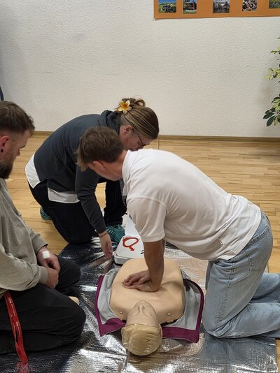 Zwei Mitarbeitende üben an einer Reanimationspuppe die Herz-Lungen-Wiederbelebung. Eine Lehrkraft überwacht die Durchführung der Herzdruckmassage während der Erste-Hilfe-Fortbildung.
