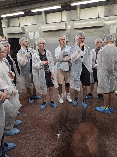 Gruppe der Azubis in Schutzkleidung mit Haarnetzen und Überschuhen in einer Lebensmittelproduktionshalle; im Hintergrund sind industrielle Anlagen und Abluftvorrichtungen zu sehen.