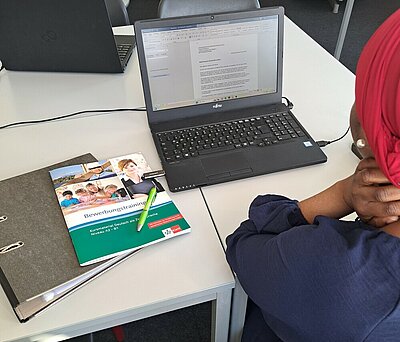 Erfolgreicher Start des Bewerbungscoachings für Migranten Eine Frau, von der man nur die Rückansicht sieht und die ein rotes Kopftuch und einen Perlenohrring trägt, sitzt an einem Tisch vor einem Laptop, auf dem man ein Bewerbungsschreiben erkennen kann. Im Laptop steckt ein USB-Stick. Neben dem Laptop liegt ein geschlossener Ordner und darauf ein Kuli sowie ein DIN A4-Heft aus dem Klett-Verlag mit dem Titel "Bewerbungstraining - Kursmaterial Deutsch als Zweitsprache - Niveau A2 - B1".