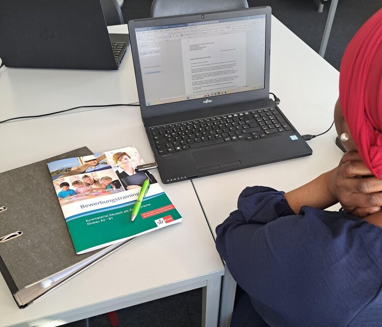 Eine Frau, von der man nur die Rückansicht sieht und die ein rotes Kopftuch und einen Perlenohrring trägt, sitzt an einem Tisch vor einem Laptop, auf dem man ein Bewerbungsschreiben erkennen kann. Im Laptop steckt ein USB-Stick. Neben dem Laptop liegt ein geschlossener Ordner und darauf ein Kuli sowie ein DIN A4-Heft aus dem Klett-Verlag mit dem Titel "Bewerbungstraining - Kursmaterial Deutsch als Zweitsprache - Niveau A2 - B1".