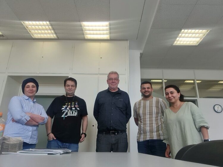 In einem Klassenzimmer stehen fünf Personen für ein Gruppenfoto bereit. In der Mitte drei Männer, außen zwei Frauen. Im Hintergrund hängt eine Uhr an der Wand, auf der 12:35 Uhr angezeigt wird. Die Neonröhren an der Decke sind angeschaltet.