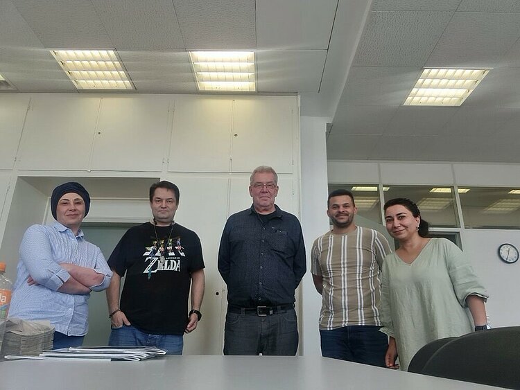 START NOW: mit Unterstützung ins Arbeitsleben In einem Klassenzimmer stehen fünf Personen für ein Gruppenfoto bereit. In der Mitte drei Männer, außen zwei Frauen. Im Hintergrund hängt eine Uhr an der Wand, auf der 12:35 Uhr angezeigt wird. Die Neonröhren an der Decke sind angeschaltet.