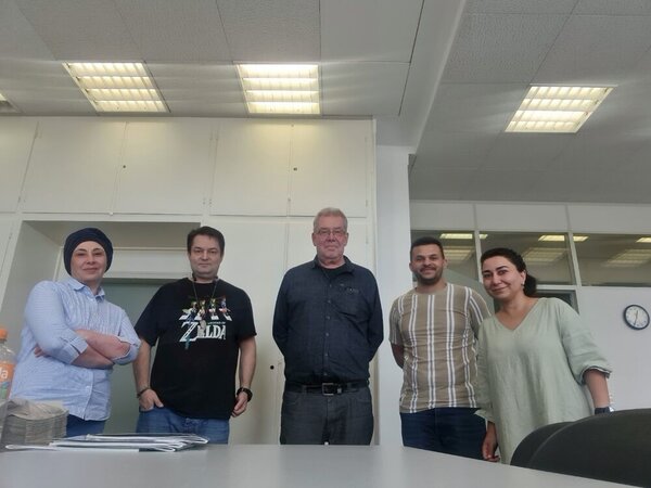 In einem Klassenzimmer stehen fünf Personen für ein Gruppenfoto bereit. In der Mitte drei Männer, außen zwei Frauen. Im Hintergrund hängt eine Uhr an der Wand, auf der 12:35 Uhr angezeigt wird. Die Neonröhren an der Decke sind angeschaltet.