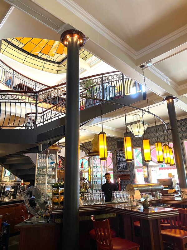 Innenansicht einer Bar in einem historischen Gebäude mit hohen Decken und Säulen. Hinter der Bar stehen zwei Barkeeper. Eine spiralförmige Treppe führt zu einer oberen Etage. Die Decke ist mit einem gelben, geometrischen Muster verziert und mehrere dekorative Lampen hängen von der Decke. Der Raum ist mit Holzmöbeln ausgestattet.