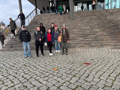 Gruppenfoto vor einer breiten Steintreppe am Veranstaltungsort: sieben Teilnehmende in Winterjacken posieren, weitere Besucher*innen stehen auf den Stufen im Hintergrund.