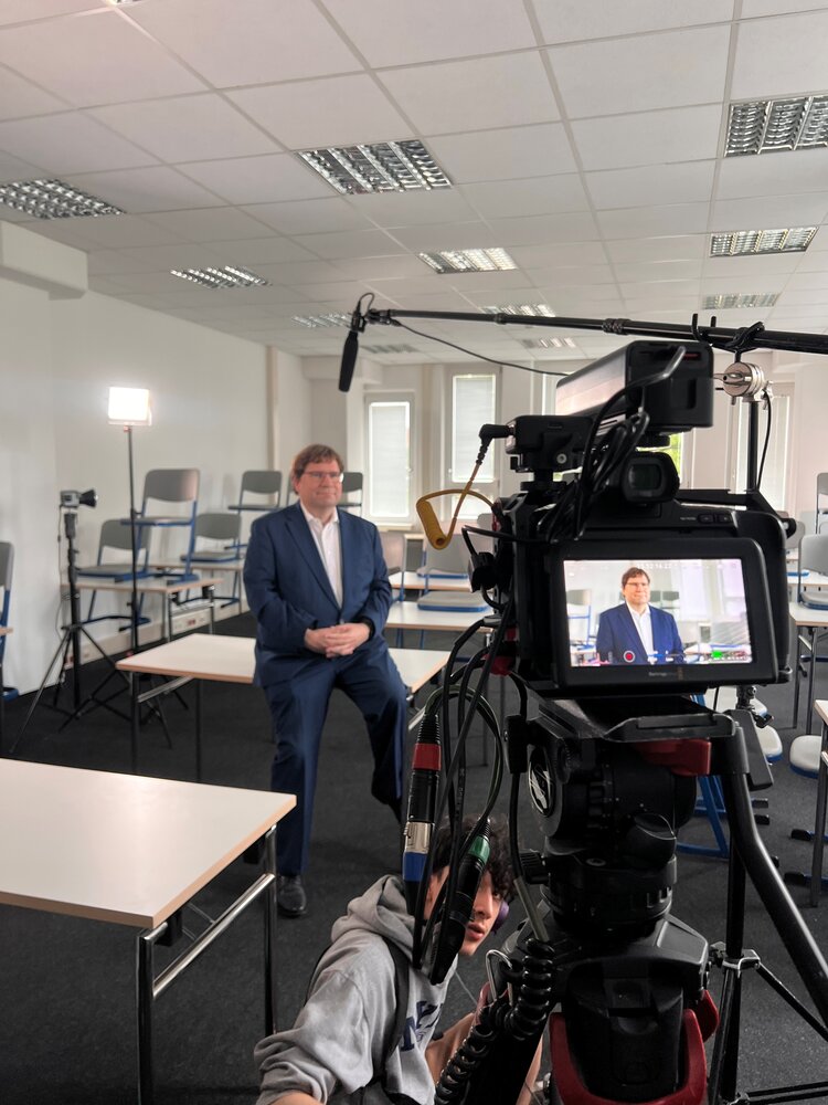 Ein Mann im Anzug sitzt während eines Videodrehs in einem Unterrichtsraum vor der Kamera; im Vordergrund sind Kameraequipment und ein Mikrofon zu sehen.