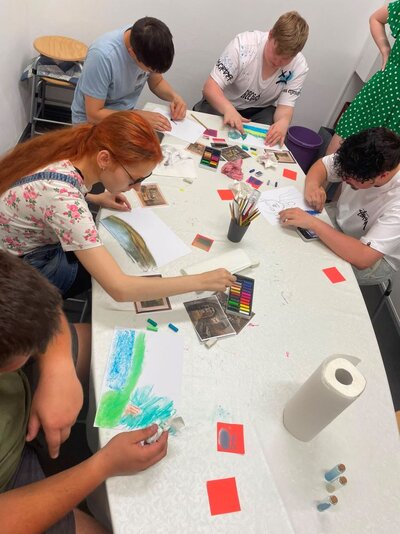 Eine Gruppe von fünf jungen Erwachsenen sitzt an einem großen, mit Papier und Malutensilien bedeckten Tisch. Sie malen und zeichnen konzentriert, während verschiedene Kunstmaterialien wie Buntstifte, Pinsel, Wasserfarben und Vorlagenbilder auf dem Tisch verstreut liegen. Die Atmosphäre wirkt kreativ und kollaborativ.