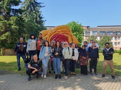 Eine Gruppe von Menschen posiert für ein Foto vor einer großen, farbenfrohen Skulptur, die wie eine überdimensionale Rose aussieht. Die Gruppe steht auf einem gepflasterten Weg in einem grünen, parkähnlichen Bereich mit Bäumen und Gebäuden im Hintergrund.