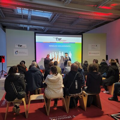 Publikum sitzt auf Hockern vor einer Bühne; auf der großen Leinwand steht ‚Herzlich Willkommen‘, zwei Personen sprechen vorne bei einer Veranstaltung.