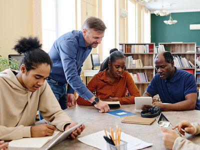 Vier Personen sitzen um einen Tisch in einem Raum, der wie ein Klassenzimmer oder ein Besprechungsraum aussieht. Eine Person steht und beugt sich über den Tisch, um den anderen etwas zu erklären oder zu zeigen. Die Personen scheinen in eine Diskussion oder eine Gruppenarbeit vertieft zu sein. Auf dem Tisch liegen verschiedene Gegenstände wie Tablets, Notizbücher und Stifte. Im Hintergrund sind Bücherregale und Pflanzen zu sehen, was auf eine Lern- oder Arbeitsumgebung hinweist.