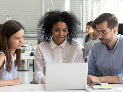 Drei Personen sitzen an einem Tisch und arbeiten gemeinsam an einem Laptop. Die Person in der Mitte, eine Frau mit lockigem Haar und einem weißen Hemd, lächelt und zeigt auf den Bildschirm. Links von ihr sitzt eine Frau mit langen dunklen Haaren und einem weißen Oberteil, und rechts ein Mann mit Brille und einem blauen Hemd, der ebenfalls auf den Laptop schaut.