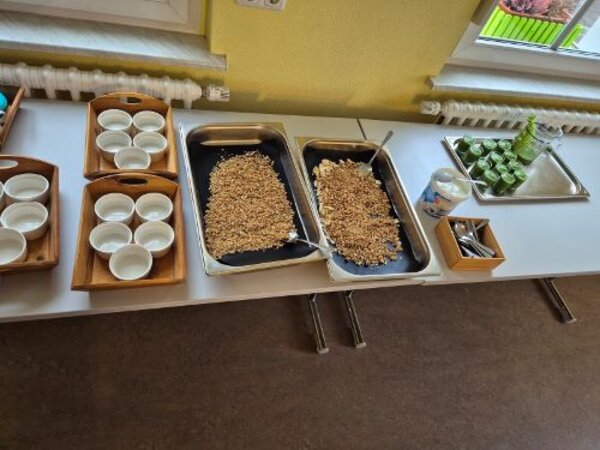 Auf einem Tisch stehen große Schalen mit Müsli, ein Becher mit Joghurt, ein Tablett mit kleinen Gläschen und einer Kanne, die mit grünem Smoothie gefüllt sind, eine Box mit Teelöffeln sowie drei Holztabletts mit weißen Müslischalen.