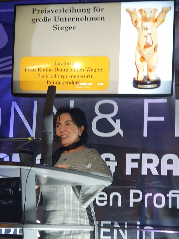 Ein Bühne mit grauem Rednerpult, an dem eine junge schwarzhaarige Frau steht, und ein Bildschirm im Hintergrund mit der Aufschrift Ausbildungsbuddy 2024.