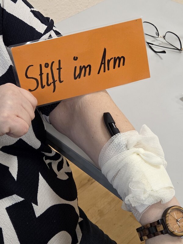 Eine Mitarbeiterin in schwarz-weiß gemusterter Bluse zeigt eine simulierte Verletzung mit einem „Stift im Arm“ und passender Verbandtechnik. Das Foto stammt aus einer praktischen Übung während einer Erste-Hilfe-Fortbildung.
