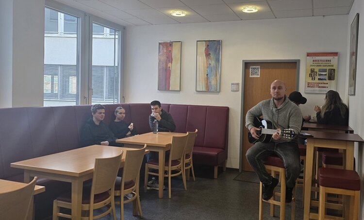 Ein Raum mit mehreren Holztischen und passenden Stühlen, an den Wänden rote Polsterbänke. Drei Teilnehmende der Euro-Schulen Dresden sitzen an einem Tisch, ein Lehrer spielt Gitarre auf einer Bank. Im Hintergrund hängen drei abstrakte, farbige Gemälde an der Wand. Rechts neben der Tür befindet sich ein Plakat mit Text und Symbolen. Große Fenster lassen Tageslicht herein.