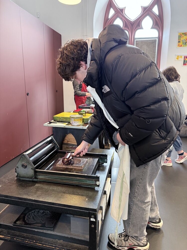 Ein junger Teilnehmender mit dunkler, wattierter Jacke und grauer Hose steht an einer Druckmaschine. Er hält ein Werkzeug und arbeitet an einer Platte, die auf dem Tisch der Maschine liegt. Im Hintergrund sind weitere Menschen und ein hoher, gotischer Fensterbogen zu sehen. Auf dem Tisch befinden sich verschiedene Behälter und Utensilien.