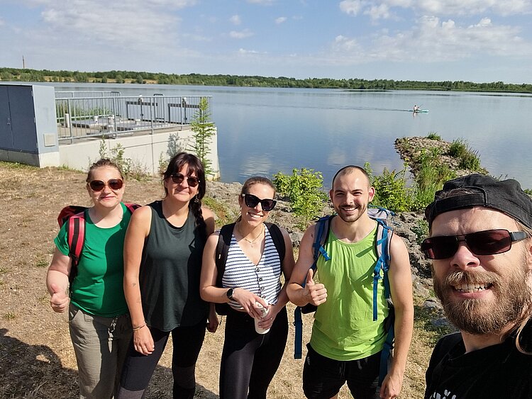 BGM-Sommerferienprogramm an ES und EA Leipzig 2025 Fünf Personen stehen am Ufer eines Sees und lächeln in die Kamera. Im Hintergrund ist Wasser mit einem Kajakfahrer sowie bewachsene Uferbereiche zu sehen.