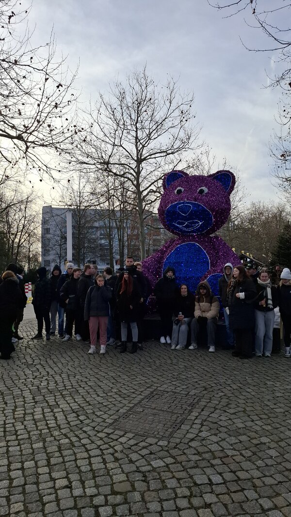 Eine größere Gruppe von Menschen steht auf einem gepflasterten Platz vor einer großen, beleuchteten Bärenfigur im Freien, umgeben von kahlen Bäumen.