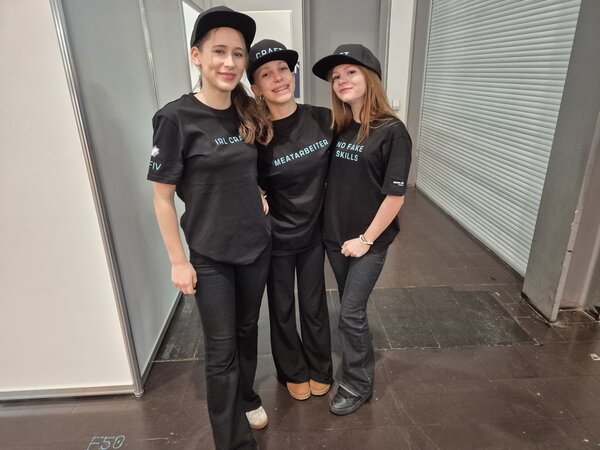 Drei lächelnde Schülerinnen mit schwarzen Kappen und schwarzen T-Shirts, auf denen "Meatarbeiter", "IRL Craft" und "No Fake Skills" steht.