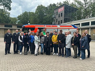 Sie haben sich für eine Gruppenfoto in Position gebracht: Mehrere Männer und Frauen stehen vor einem Einsatzfahrzeug der Berliner Feuerwehr, sie sind umrahmt von Feuerwehrleuten in ihrer Ausgehuniform. Hinter dem Fahrzeug kann man einen großen Turm erkennen, an dem das Logo der Berliner Feuerwehr- und Rettungsdienst-Akademie (BFRA) prangt.
