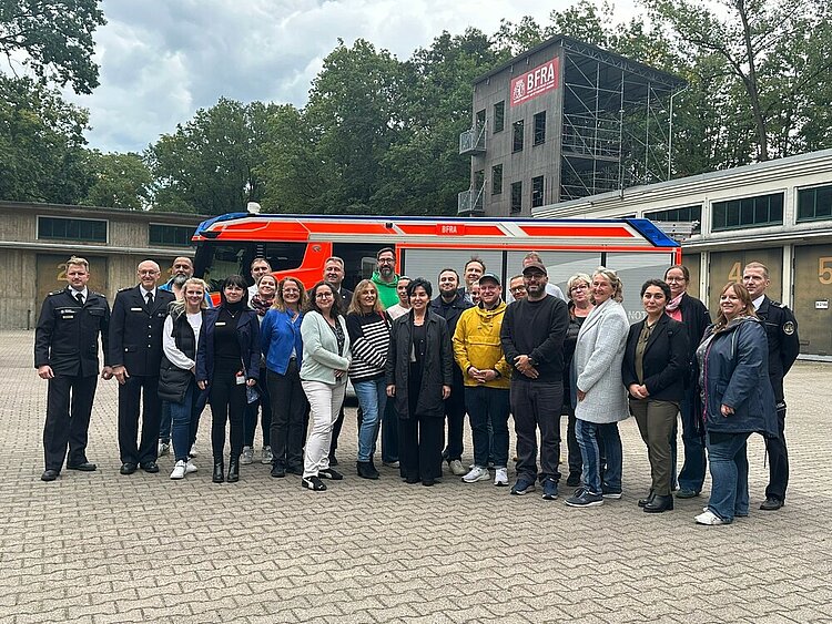 Sie haben sich für eine Gruppenfoto in Position gebracht: Mehrere Männer und Frauen stehen vor einem Einsatzfahrzeug der Berliner Feuerwehr, sie sind umrahmt von Feuerwehrleuten in ihrer Ausgehuniform. Hinter dem Fahrzeug kann man einen großen Turm erkennen, an dem das Logo der Berliner Feuerwehr- und Rettungsdienst-Akademie (BFRA) prangt.