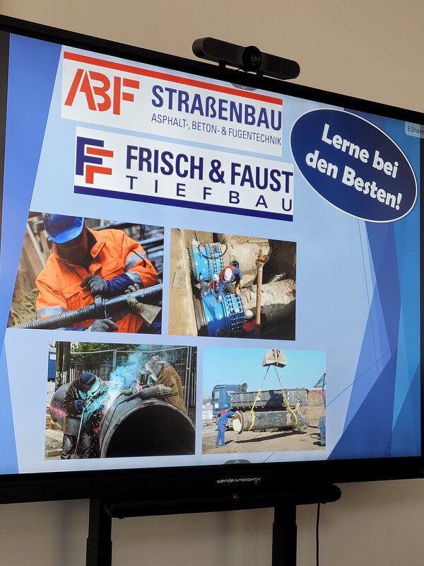 Präsentation auf einem Bildschirm über die Unternehmen ABF Straßenbau und Frisch & Faust Tiefbau. Zu sehen sind mehrere Fotos von Arbeiten im Tief- und Rohrleitungsbau sowie der Schriftzug „Lerne bei den Besten!“.