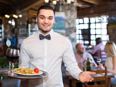 Ein Kellner in einem weißen Hemd und mit einer Fliege steht lächelnd in einem Restaurant. Er hält ein Tablett mit einem Teller frischem Salat, der mit Tomaten und anderen Gemüsesorten garniert ist. Auf dem Tablett steht außerdem ein Glas Rotwein. Der Kellner wirkt freundlich und einladend, während er den Arm ausstreckt, als würde er den Gast zum Tisch begleiten. Im Hintergrund sind Gäste zu sehen, die an Tischen sitzen und essen. Das Restaurant hat eine gemütliche Atmosphäre mit rustikalen Holzmöbeln und warmen Lichtern.