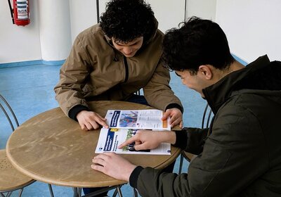 Auf dem Bild sitzen zwei Jugendliche mit Migrationshintergrund an einem kleinen, runden Holztisch im Gebäude der Euro-Schulen Leipzig. Sie beugen sich über ein farbiges Informationsheft des Projekts ‚KarriereStart III‘ und zeigen mit den Fingern auf einzelne Inhalte. Beide tragen wetterfeste Jacken und wirken konzentriert, während sie sich über berufliche Orientierungsmöglichkeiten austauschen. Der Raum hat einen hellen Boden, weiße Wände und Metallstühle. Die Szene vermittelt Teamarbeit, Beratungsatmosphäre und aktive Teilnahme an der beruflichen Zukunftsplanung.
