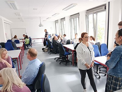 Job-Speed-Dating vom 22.07.2025 ein voller Erfolg! Ein heller Seminarraum mit mehreren Gruppen von Menschen, die an Tischen sitzen und diskutieren. Im Vordergrund stehen zwei Frauen im Gespräch, im Hintergrund arbeiten Teilnehmende in Kleingruppen an Aufgaben.