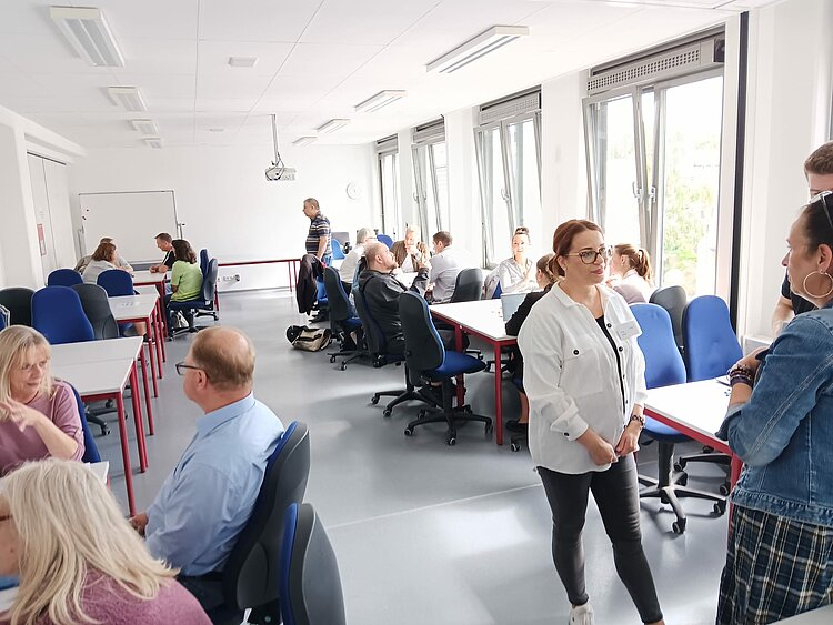 Job-Speed-Dating vom 22.07.2025 ein voller Erfolg! Ein heller Seminarraum mit mehreren Gruppen von Menschen, die an Tischen sitzen und diskutieren. Im Vordergrund stehen zwei Frauen im Gespräch, im Hintergrund arbeiten Teilnehmende in Kleingruppen an Aufgaben.