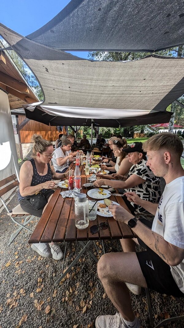 Eine Gruppe von Männern und Frauen in sommerlicher Kleidung sitzt an einer langen Tafel von Gartentischen und isst und trinkt. Es stehen verschiedene Getränkeflaschen und Gläser auf dem Tisch. Über den Tisch sind Tücher gespannt.