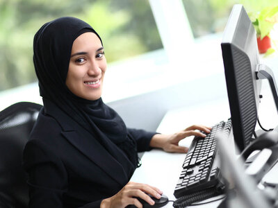 Eine Frau mit einem schwarzen Hijab sitzt an einem Schreibtisch in einem Büro. Sie trägt ein schwarzes Jackett und arbeitet an einem Computer. Sie lächelt und scheint konzentriert oder zufrieden zu sein. Auf dem Schreibtisch vor ihr befinden sich eine Tastatur und eine Maus. Im Hintergrund ist ein Fenster zu sehen, das Tageslicht hereinlässt und eine grüne Landschaft zeigt.